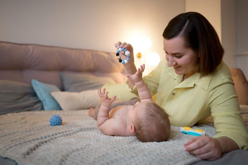 A parent and baby interact on a cozy bed, with colorful toys scattered around in a warmly lit, inviting room.