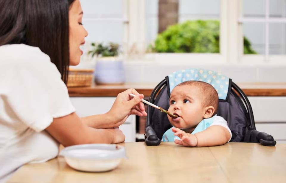A caregiver feeds a seated baby in a high chair, with a bowl of food on the table, in a bright kitchen setting.