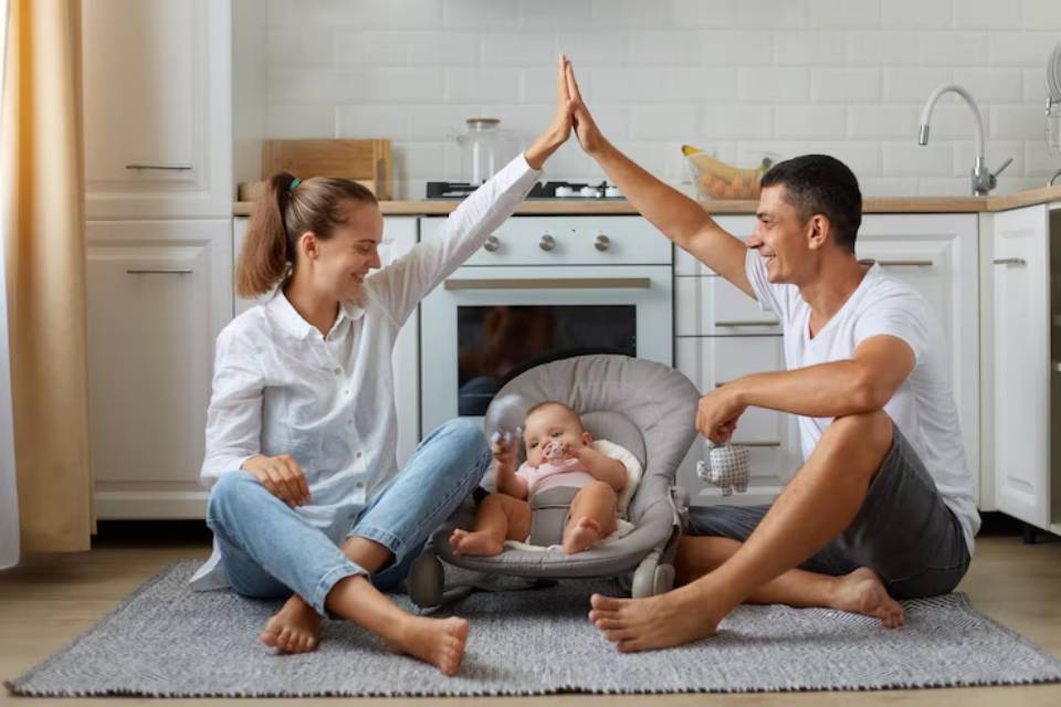A couple sitting on a rug in a kitchen, playfully raising their hands to each other, with a baby in a seat nearby.