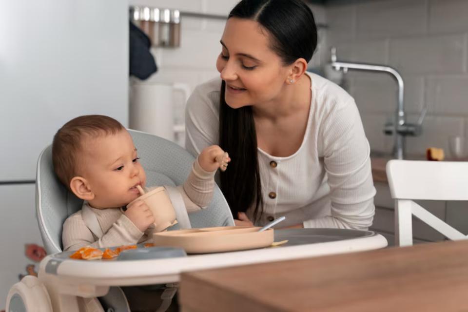  A caregiver interacts with a young child seated in a high chair during a meal, surrounded by a cozy kitchen setting.