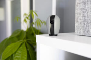 A modern security camera in gray and black sits on a white shelf next to a plant, with a textured gray box in the background.