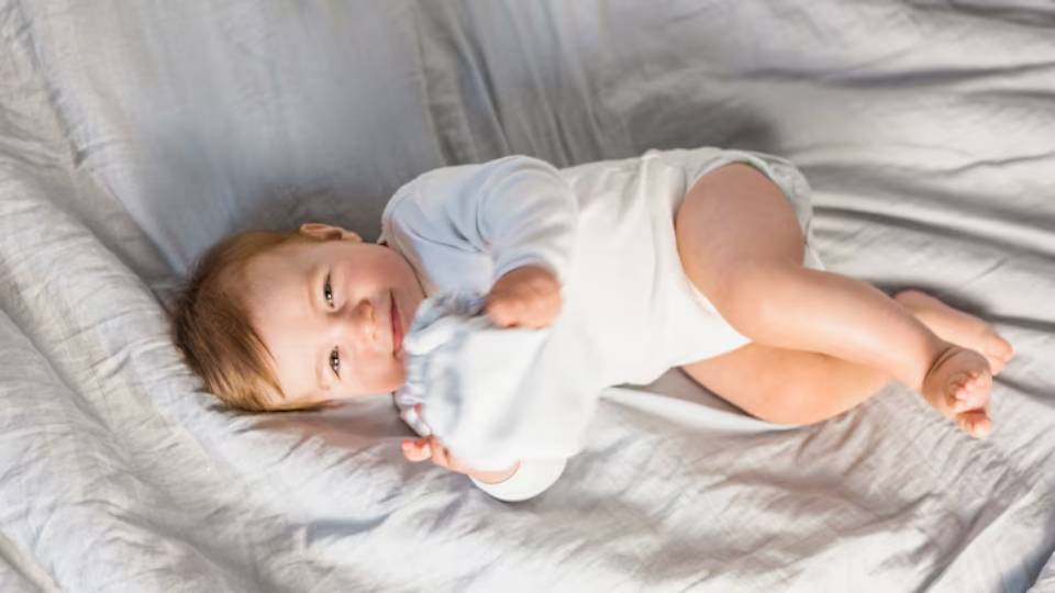 A baby in a white onesie lies on a soft, light grey blanket, curled up comfortably with chubby limbs.