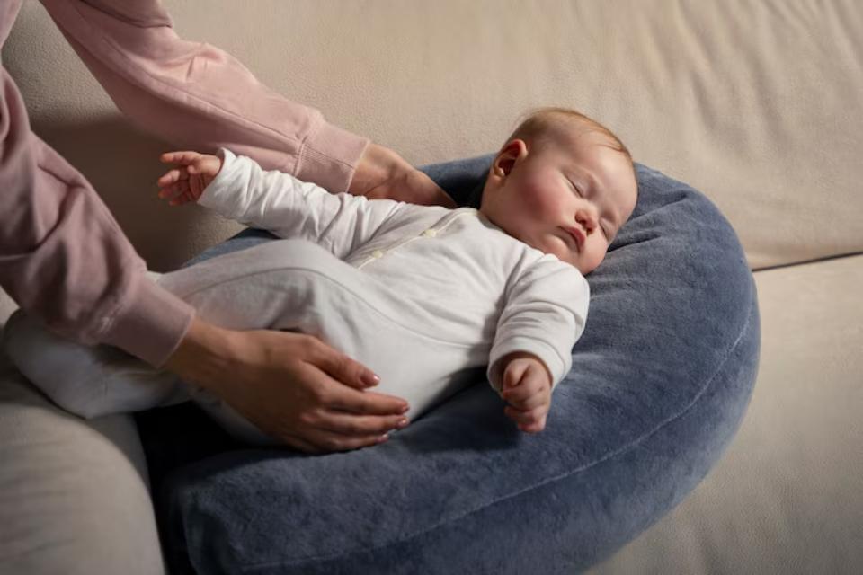 A baby in a white onesie is gently cradled by a person wearing a pink sweater, resting on a soft, round blue pillow.