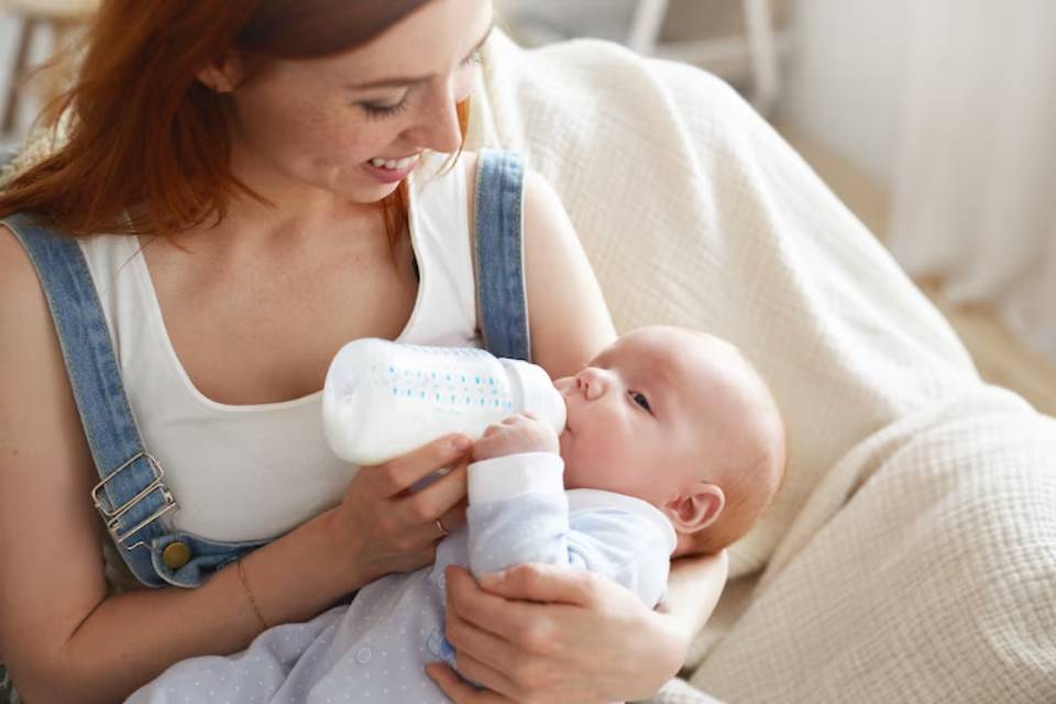 A woman in a white tank top and denim overalls gently feeds a baby from a bottle while sitting on a cozy blanket.