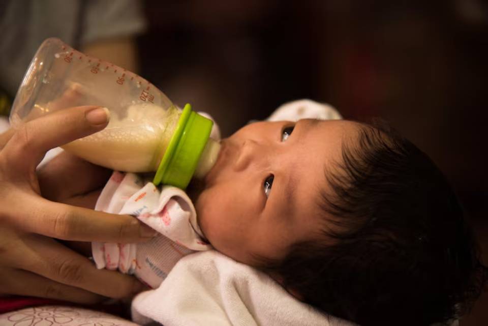 A hand holds a baby bottle filled with milk, as a baby is cradled in a soft blanket, creating a nurturing feeding moment.