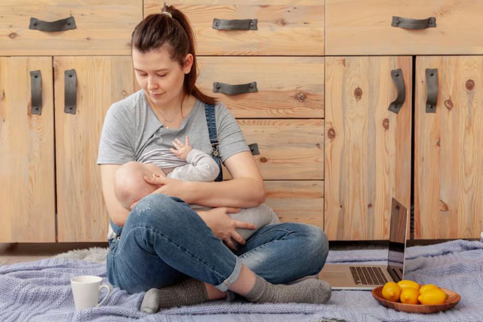  A woman in casual clothing, sitting cross-legged on a rug, breastfeeds a baby while using a laptop and has a cup of tea nearby.