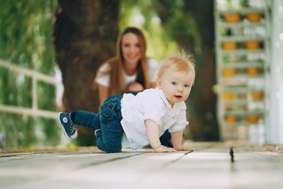  A baby with blonde hair crawls on a wooden pathway surrounded by greenery, with a figure in the background.