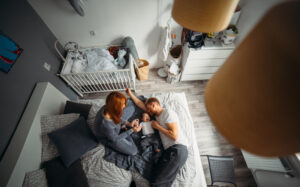 A couple relaxes on a bed, tenderly holding a small child, surrounded by cozy linens in a warmly decorated nursery.