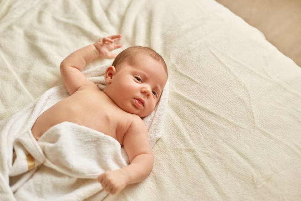 A peaceful baby lies on a soft, light-colored blanket, partially wrapped in a white towel, with one arm reaching out playfully.