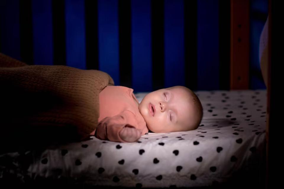  A peaceful baby resting in a crib, covered by a warm blanket, on a heart-patterned sheet, against a dark blue backdrop.