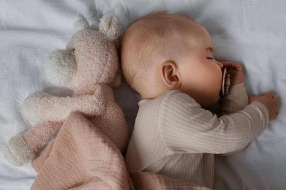 Newborn baby sleeping peacefully on a bed next to a soft plush teddy bear, wrapped in a beige blanket.