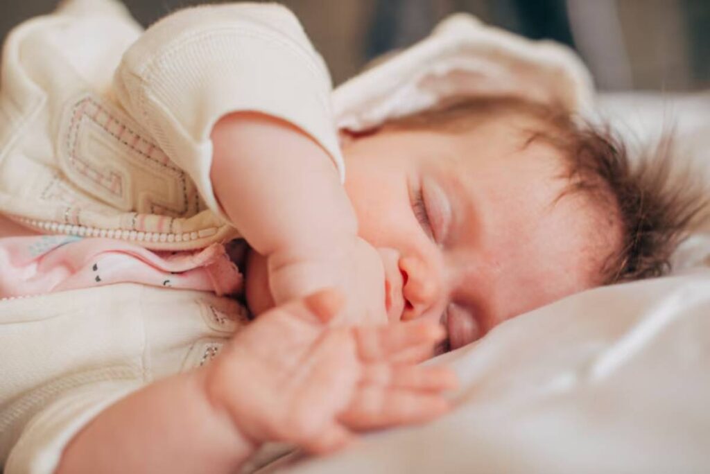 A close-up of a baby resting on a soft surface, wearing a light sweater and pink onesie, with tiny hands gently stretched out.