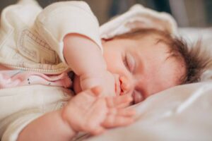 A close-up of a baby resting on a soft surface, wearing a light sweater and pink onesie, with tiny hands gently stretched out.