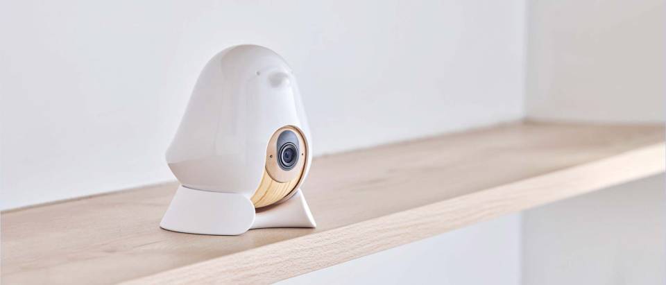 A stylish, white security camera shaped like a bird, with a wooden accent, perched on a light wooden shelf against a minimalistic background.
