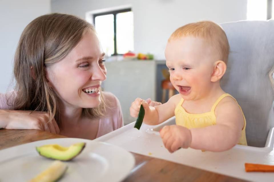 A toddler in a high chair explores a cucumber slice while seated at a wooden table with avocado and carrot on a plate.