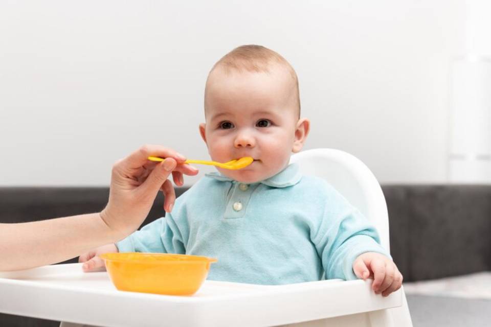 A baby in a high chair is being fed with a yellow spoon by a caregiver, accompanied by a bowl of food on the tray.