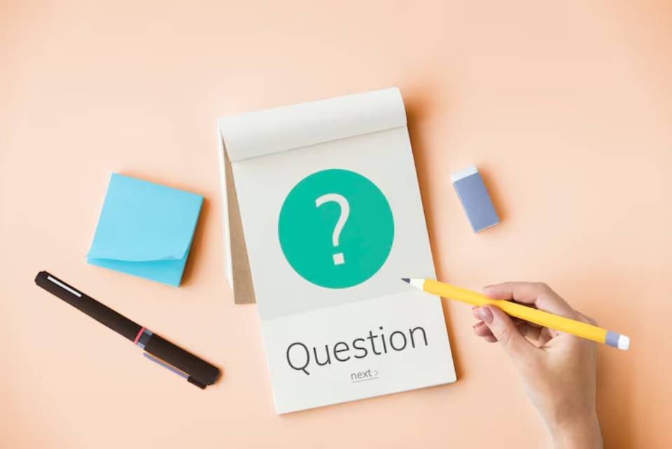 A hand holding a yellow pencil above a notepad displaying a green question mark, surrounded by post-it notes and stationery on a peach background.