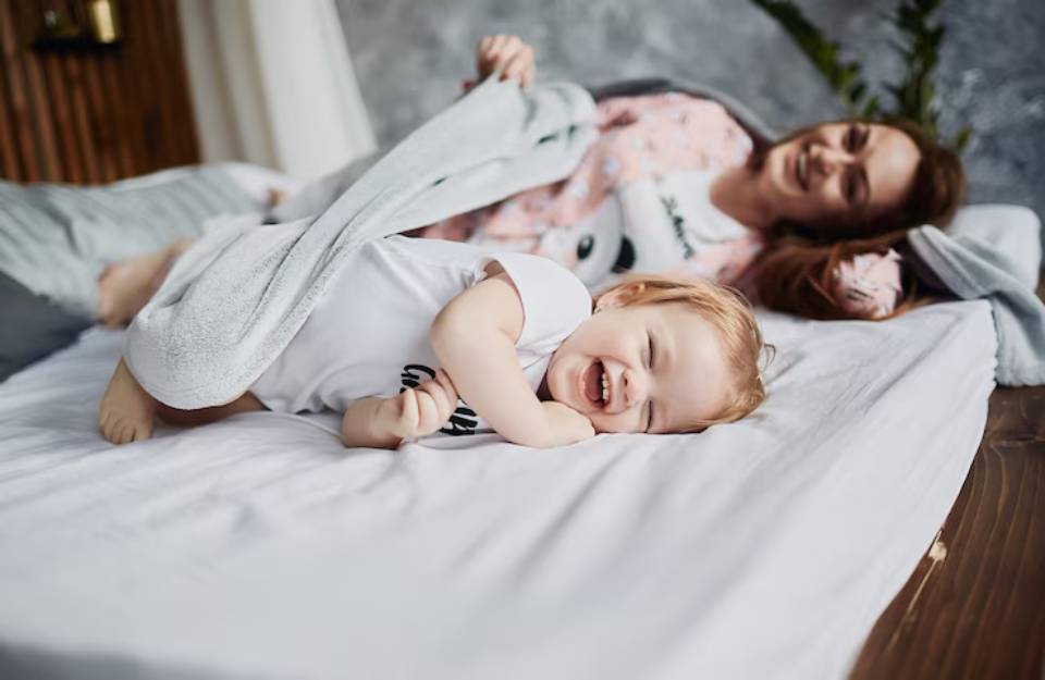 A cozy scene of a child in a white shirt and another person relaxing on a bed, partially covered with a gray blanket.