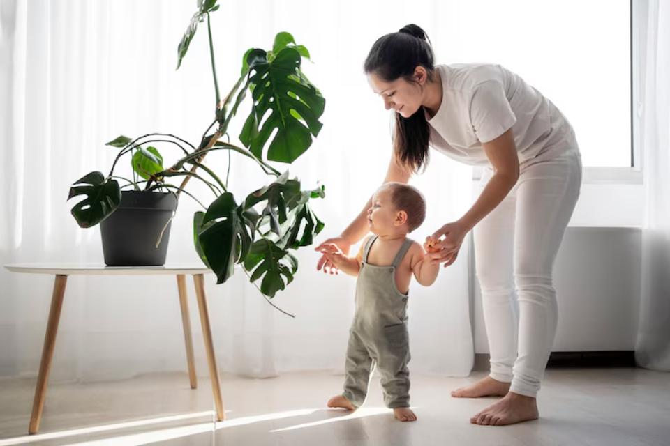  A woman gently guides a toddler, standing in a bright room with a large potted plant on a white table, sunlight streaming through the window.