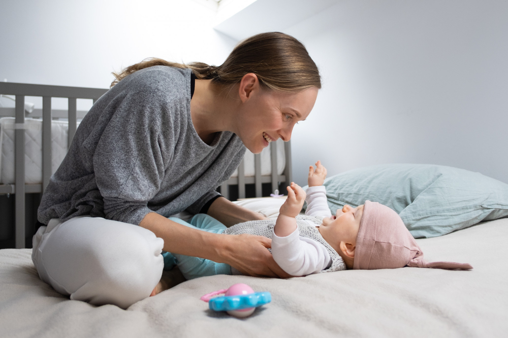  A woman in a cozy outfit interacts with a baby on a bed surrounded by soft pillows and a crib in a bright, serene room.