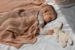 A peaceful baby sleeps on a white bed, wrapped in a soft blush blanket, with a plush bunny toy beside them.