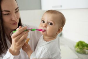 A caregiver is gently feeding a baby with a green and pink spoon in a bright kitchen setting, with fresh vegetables in the background.