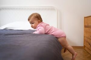 A baby in a pink onesie is crawling over the edge of a bed with a textured dark blanket, in a bright, minimalistic room.