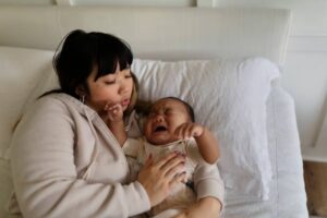 A mother and her baby relax together on a white bed, sharing a tender moment in a cozy, softly lit room.