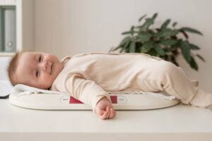 A baby in a beige outfit is lying comfortably on a white digital scale, with a green plant in the background.