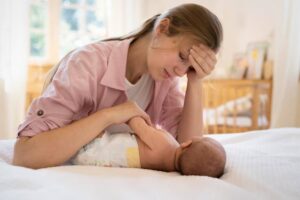 A woman in a pink shirt gently interacts with her baby on a bed in a bright, cozy room with wooden furniture.