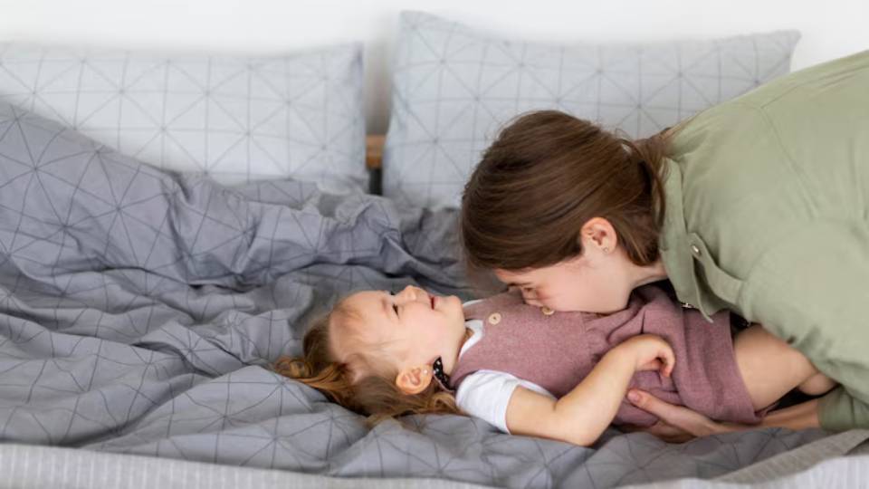 A woman leans down to lovingly interact with a child lying on a gray-patterned bed with plush blankets and pillows.