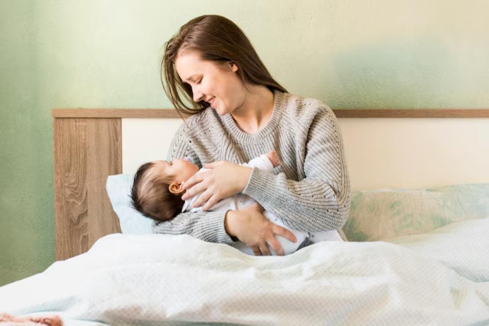  A woman gently cradles her baby in her arms while sitting on a bed, surrounded by soft blankets and a calming green wall.