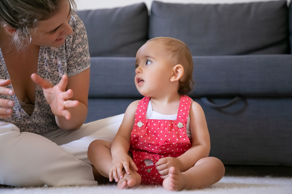 A child in a red polka dot outfit sits on a soft rug, listening attentively while an adult gestures animatedly nearby.