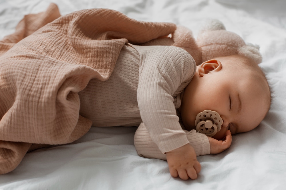 A peaceful baby sleeps on a soft white bed, wrapped in a peach blanket, with a pacifier in mouth and a teddy bear nearby.