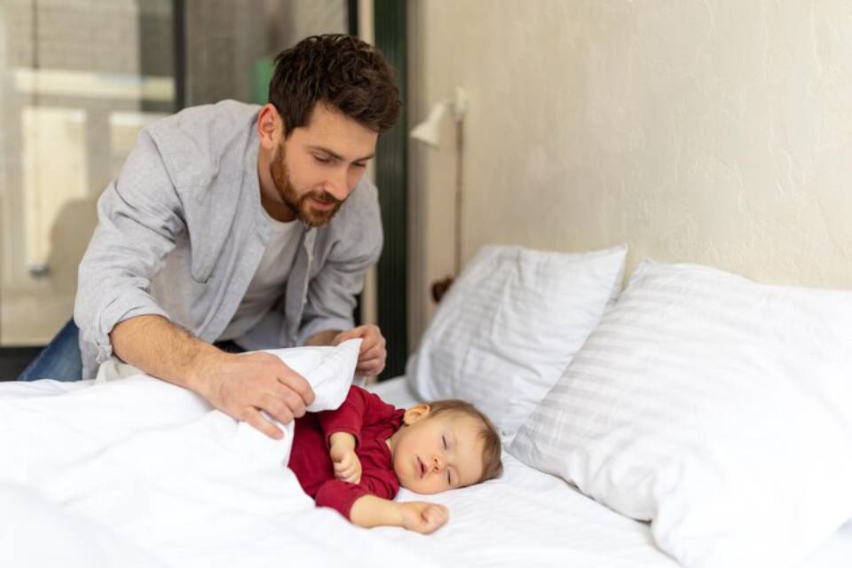 A caregiver gently tucks a child into a bed with white linens in a cozy, well-lit room.