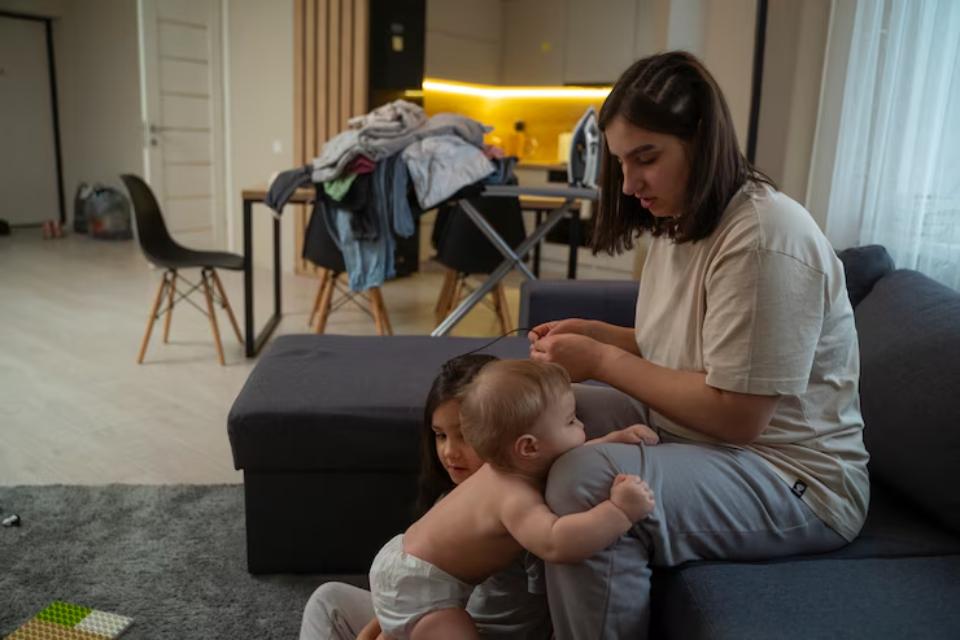  A woman sits on a couch, attending to two small children, with a cluttered dining area and warm lighting in a modern living room.