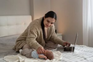 A person in a cozy sweater works on a laptop while gently interacting with a baby lying on a bed draped in a light blanket.