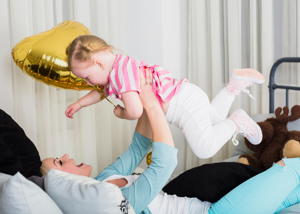 A child in a pink striped shirt is playfully lifted by an adult while holding a gold heart-shaped balloon, with cozy pillows in the background.