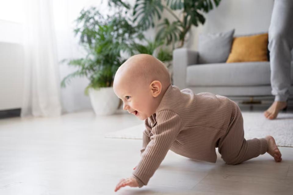  A baby in a beige outfit crawls on a light wooden floor, with green plants and a gray couch in the background.