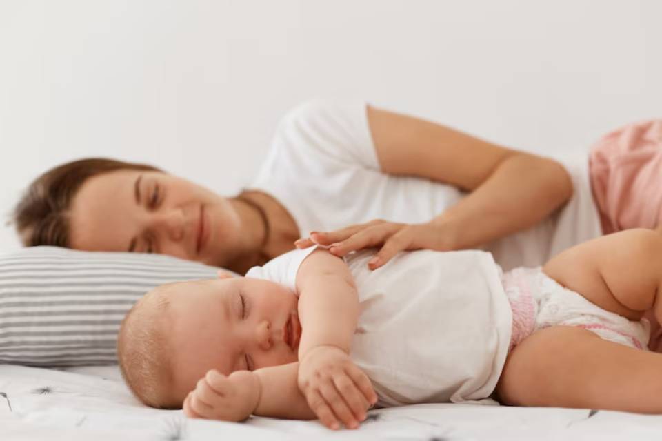 A mother lies on a bed, smiling at her baby, as they share a tender moment together in a soft, light-filled room.