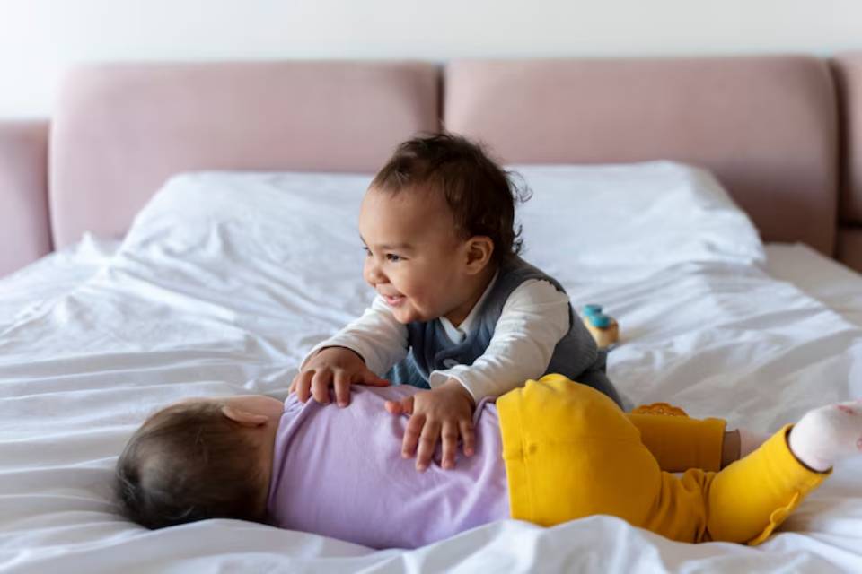 Two babies play on a white bed, one lies on their stomach while the other gently reaches out to touch them.