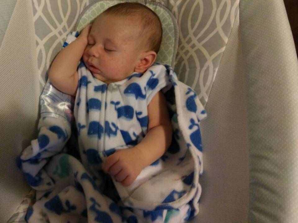 A cozy infant is relaxing in a soft, whale-patterned blanket, one hand resting on their forehead while nestled in a baby seat.