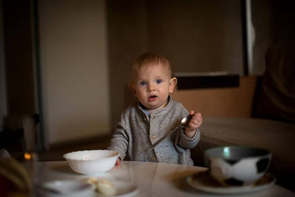 A toddler in a cozy sweater holds a spoon, sitting at a table with a bowl and a cup, in a softly lit interior.