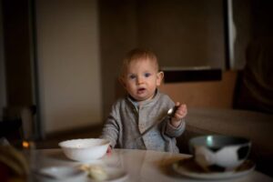 A toddler in a cozy sweater holds a spoon, sitting at a table with a bowl and a cup, in a softly lit interior.