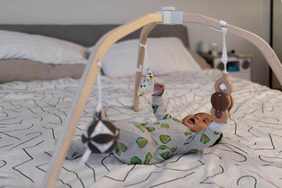A baby in a green avocado-patterned outfit plays beneath a wooden activity gym, surrounded by soft bedding and pillows.