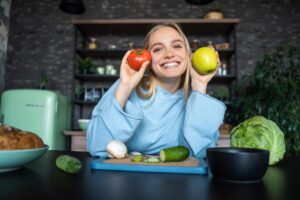 A person holds a red tomato and a green apple, surrounded by fresh vegetables and bread on a kitchen counter with a retro fridge in the background.