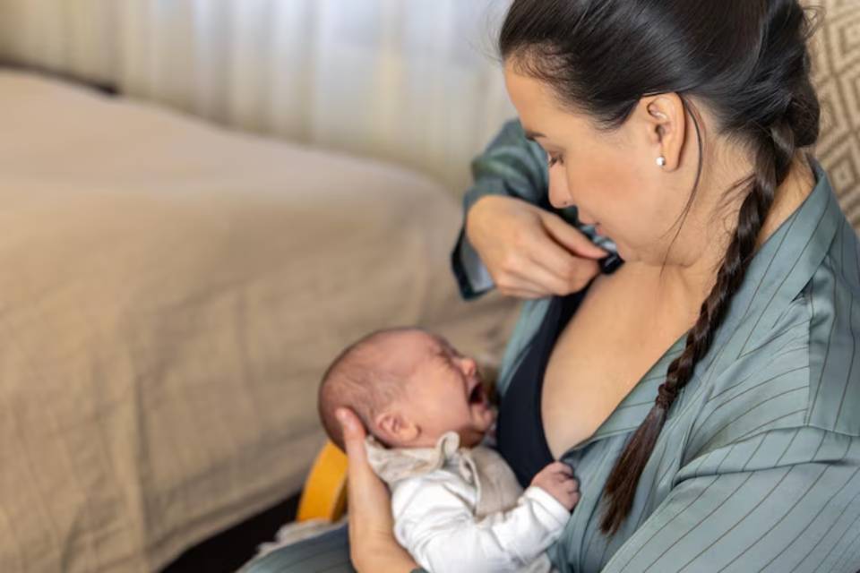 A mother cradles a crying newborn in a cozy setting, wearing a light green robe, with warm sunlight filtered through the curtains.