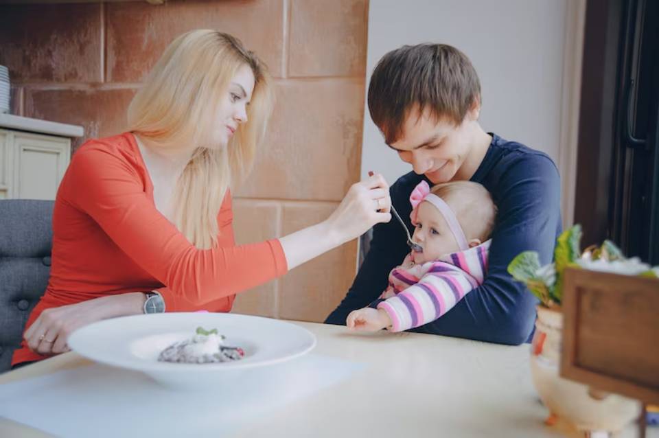 A woman in a red top feeds a baby in a pink-striped outfit while a man watches, with a dish of food on the table.