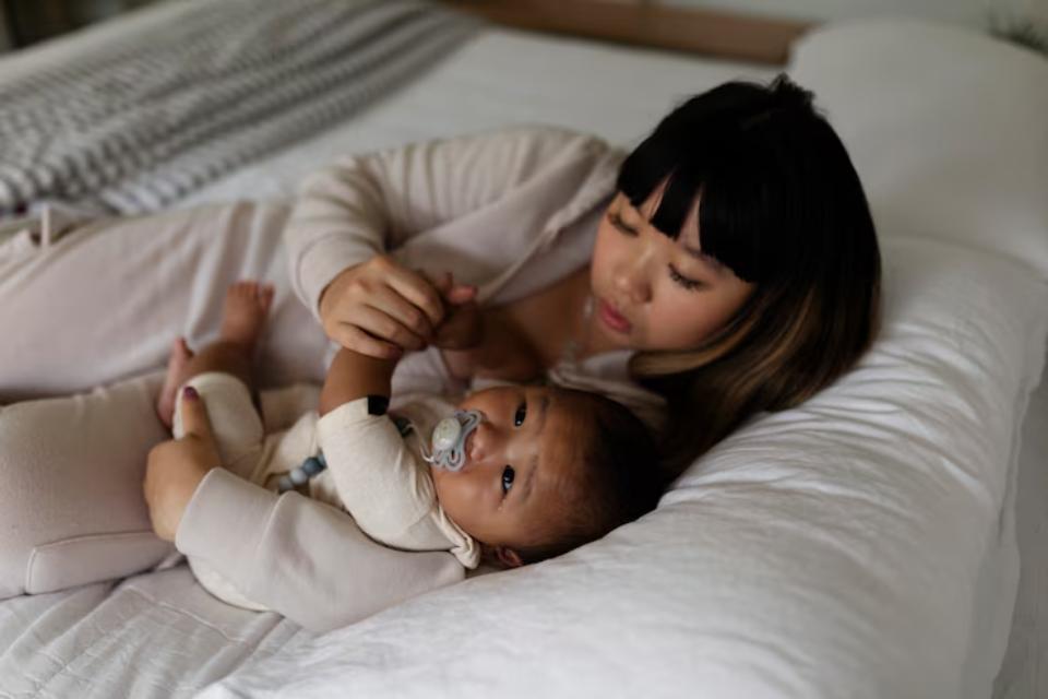  A woman in a light-colored outfit gently holds her baby's hand while lying on a cozy bed, surrounded by soft bedding and warm light.