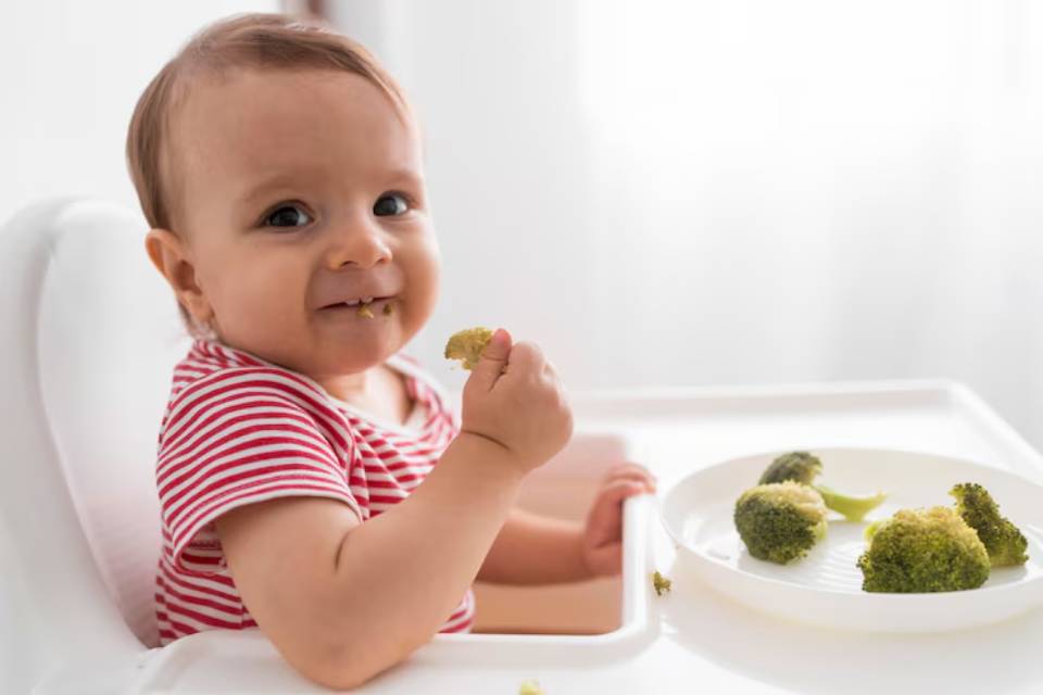 A toddler in a high chair wears a striped shirt and holds a piece of broccoli, with more broccoli arranged on a plate nearby.